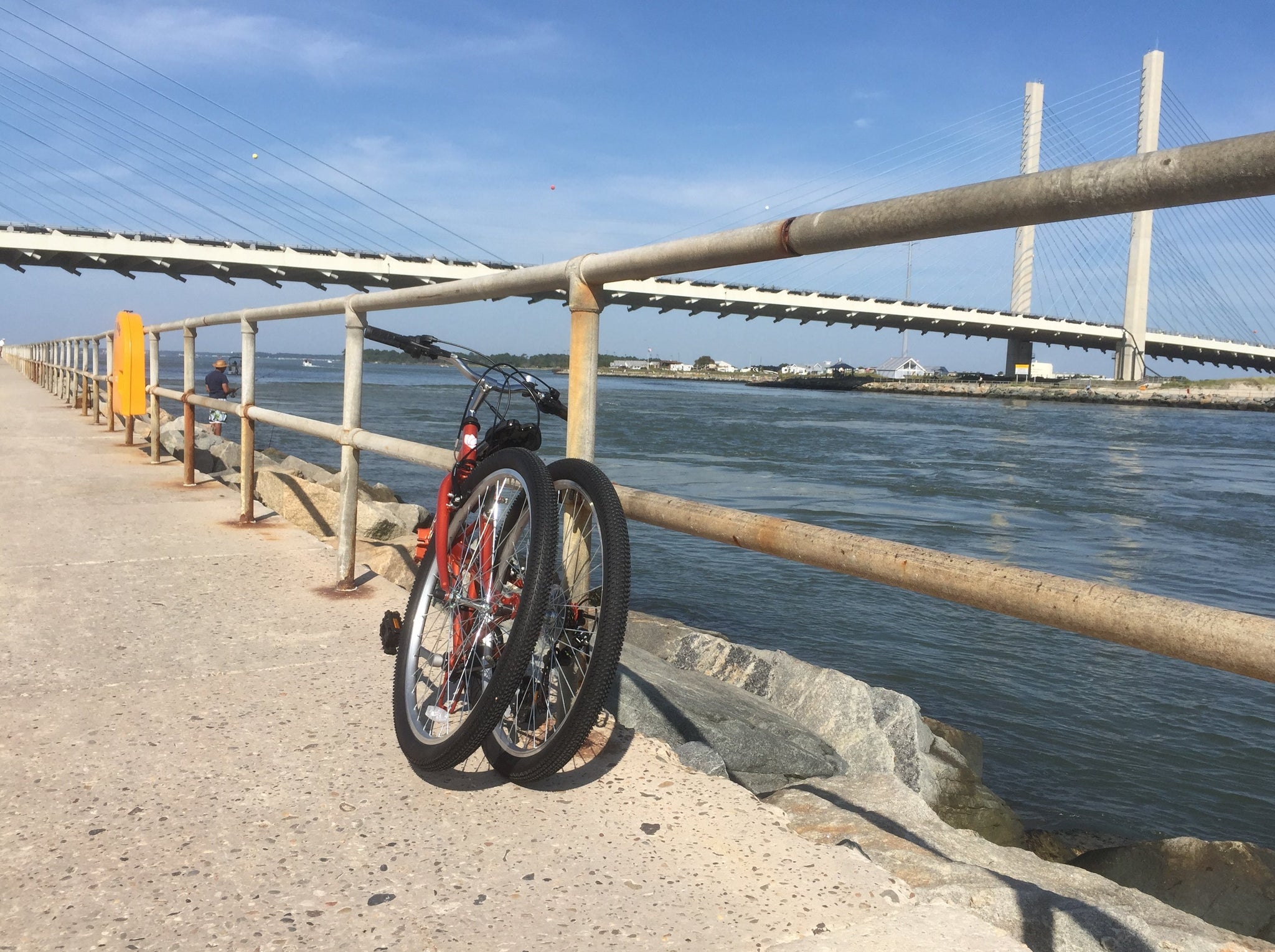 Orange colored folding bike alongside a body of water with bridge in the background.
