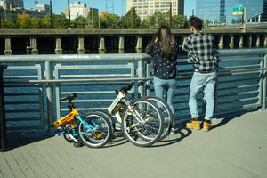 Girl and boy looking off into the distance with two folding bikes beside them on a boardwalk by a body of water.
