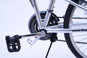 Foldable bike pedal attached to crank arm. The image also depicts a front derailleur, bike chain ring, bike chains, a metal bike stand, and part of the wheel.